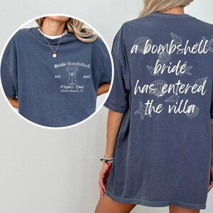 Op de afbeelding: Marineblauw T-shirt met witte tekst en schelpmotieven. De voorkant leest "Bride Bombshell" met "EST. 2025" en "Megan's Bach Miami Beach, FL". De achterkant zegt "a bombshell bride has entered the villa".