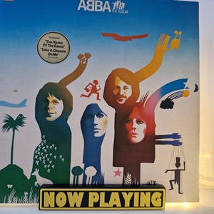 Op de afbeelding: De albumhoes van ABBA's "The Album" met de bandleden tegen een blauwe lucht. De hoes bevat de tekst "Features The Name Of The Game Take A Chance On Me". Een geel bord met de tekst "NOW PLAYING" staat voor het album.