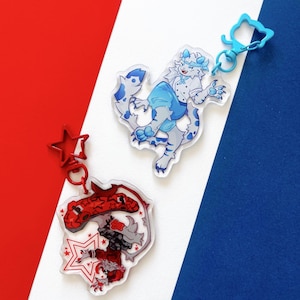 May include: Two clear acrylic keychains with cartoon character designs. One features a red and black creature with a star-shaped outline and a red star-shaped clasp. The other has a blue and white character with a blue cat-shaped clasp. The background is red, white, and blue.
