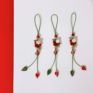 May include: Three decorative floral ornaments with red and white roses, green leaves, and red heart-shaped beads. The ornaments are suspended by green cords and are set against a white background with a red border.