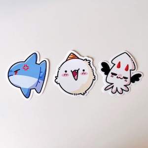 May include: Three cartoon stickers: an angry blue shark, a happy white creature with a party hat, and a cute white squid with black wings. Each sticker has a black outline and is on a white background.