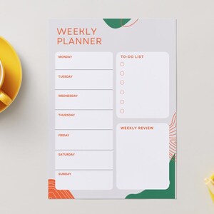 Könnte beinhalten: Ein Wochenplaner mit den Worten "WEEKLY PLANNER" in Orange. Der Planer hat Abschnitte für jeden Wochentag, eine To-Do-Liste und einen Wochenüberblick. Eine gelbe Tasse Tee und ein gelbes Tablett liegen auf dem Tisch.