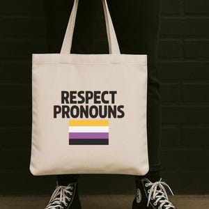 May include: A cream-colored tote bag with the words "RESPECT PRONOUNS" in black block letters. Below the text is a horizontal non-binary pride flag. The bag is held by a person wearing black pants and black high-top sneakers.
