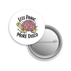 May include: A white button pin with a cartoon turtle design. The turtle has a disco ball shell in shades of pink, purple, and peach. The text reads "Less Panic More Disco" in black, stylized lettering. The pin is round and has a metal backing.
