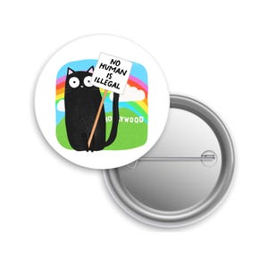 May include: A white button featuring a black cat holding a sign that reads "NO HUMAN IS ILLEGAL" against a backdrop of a rainbow, clouds, and green grass. The word "HOLLYWOOD" is at the bottom. The button is round and has a pin back.