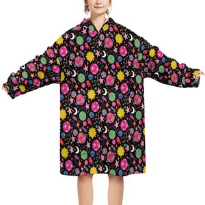 May include: A black hooded blanket with a colorful space-themed pattern. The design includes planets, stars, moons, suns, and rockets in pink, yellow, blue, and orange. The blanket has long sleeves and a large hood.