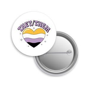 May include: A white button pin with the words "THEY/THEM" in purple lettering. The pin features a heart in the non-binary pride flag colors: yellow, white, lavender, and black. Small star shapes surround the heart.