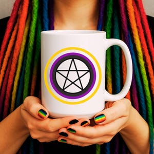 May include: White ceramic mug featuring a black pentagram design within a yellow, purple, and black circle. The mug is held by hands with rainbow-colored nail art, against a backdrop of rainbow-colored dreadlocks.