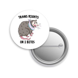 May include: A white button pin with a cartoon opossum wearing a transgender pride flag bandana. The pin has the text "TRANS RIGHTS" above the opossum and "OR I BITES" below it. The pin is round and has a metal backing.