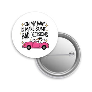 May include: A white button pin with the text "ON MY WAY TO MAKE SOME BAD DECISIONS" and a cartoon image of a swan wearing sunglasses driving a pink convertible. The pin is round and has a metal backing.
