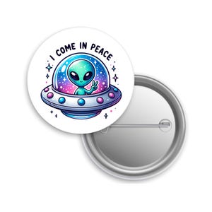 May include: A white button pin featuring a cartoon alien in a spaceship with the text "I COME IN PEACE." The alien is green with large black eyes and is giving a peace sign. The spaceship is silver, blue, and pink.