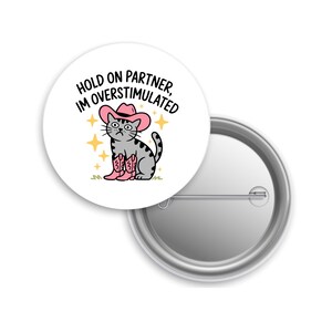 May include: A white button featuring a cartoon cat wearing a pink cowboy hat and boots. The cat has a distressed expression and is surrounded by gold stars. Text reads "HOLD ON PARTNER, IM OVERSTIMULATED."