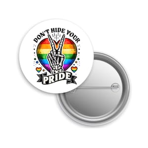 May include: A white button with a rainbow heart design and a skeleton hand making a peace sign. The text reads "DON'T HIDE YOUR PRIDE". Two small rainbow hearts are on either side of the heart.