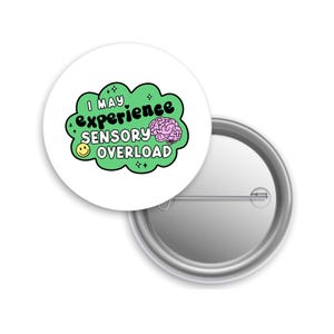 May include: A white button pin with the text "I MAY experience SENSORY OVERLOAD" in black and green, with a pink brain graphic and a yellow smiley face. The pin is round with a metal backing.