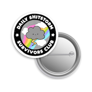 May include: A round button pin with the text "DAILY SHITSTORM SURVIVORS CLUB" in a black border. The pin features a gray cloud with a sad face, a rainbow, and a lightning bolt. The pin is white with a metal backing.