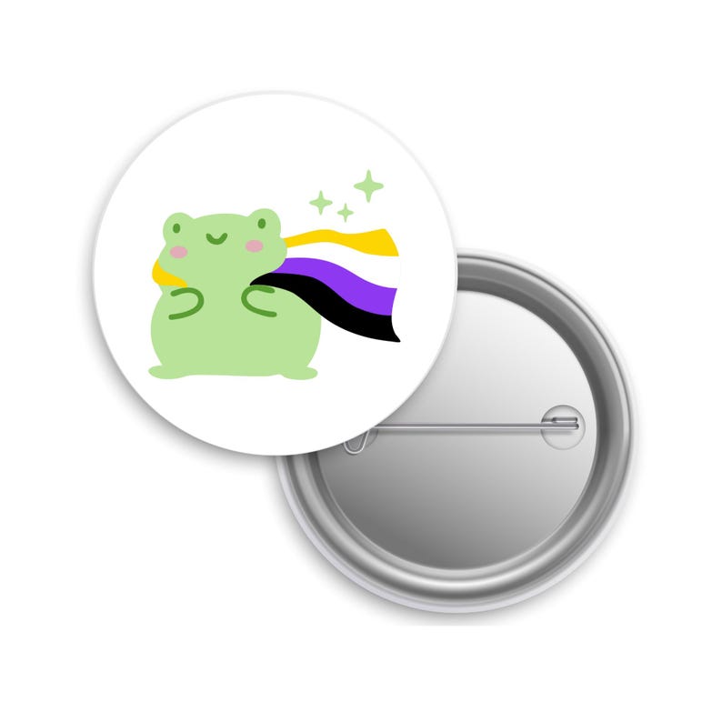 May include: A white, circular badge with a cartoon frog in light green, pink cheeks, and a non-binary pride flag. The flag is yellow, purple, black, and white. Green stars are in the background. The badge is made of metal.