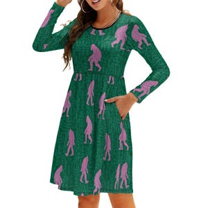 May include: A green long-sleeved dress with a pattern of pink Bigfoot figures. The dress has a round neckline, pockets, and a flared skirt. The Bigfoot figures are in various poses, creating a playful and whimsical design.