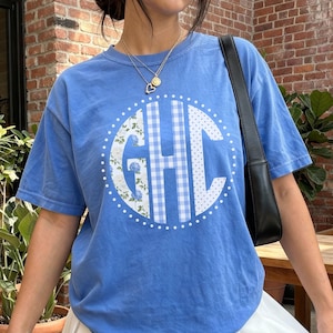 May include: A light blue t-shirt featuring a monogram design. The monogram includes the letters GHC, with a floral and gingham pattern. The shirt has short sleeves and a crew neck. The design is centered on the front of the shirt.