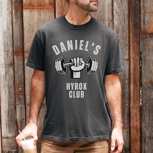 May include: Dark gray t-shirt with the text "DANIEL'S" above a graphic of a hand holding a barbell, and "HYROX CLUB" below. The shirt is short-sleeved and has a crew neck.