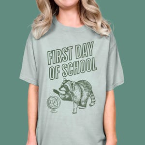 May include: A light sage green short-sleeve t-shirt featuring a dark green graphic print. The design includes the text "FIRST DAY OF SCHOOL" in a bold font above an illustration of a raccoon standing next to a small globe on a stand.