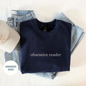 May include: Navy blue sweatshirt with the text "obsessive reader" in white, paired with light wash denim jeans and a beige baseball cap. The sweatshirt is a crew neck style. The baseball cap has the letter "N" on it.