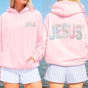 May include: A light pink hoodie with the word "JESUS" embroidered on the front and back. The letters are made of various patterned fabrics. The hoodie has a hood and a front pocket. The model is wearing blue and white striped shorts.