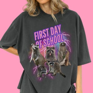 May include: A charcoal gray t-shirt featuring a graphic print of five raccoons arranged in a group. Above the raccoons, the text FIRST DAY OF SCHOOL is printed in purple letters. The design includes a purple lightning bolt background effect.