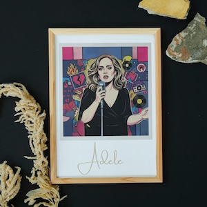 Puede incluir: Impresión artística enmarcada que presenta una ilustración estilizada de Adele cantando con un micrófono. La obra de arte tiene un fondo colorido con elementos musicales y el nombre "Adele" en escritura dorada. El marco es de madera clara.