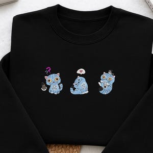 Op de afbeelding: Zwart sweatshirt met drie blauwe cartoonkatten met verschillende uitdrukkingen. De katten hebben grote ogen en worden afgebeeld met denkballonnen en accessoires. Het sweatshirt is gemaakt van een zacht materiaal en is geschikt voor vrijetijdskleding.