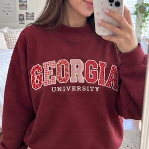 May include: A maroon crewneck sweatshirt featuring the word GEORGIA in large, block-style letters. Each letter has a unique red and white pattern, including stars, gingham, floral, stripes, and polka dots. The word UNIVERSITY is printed below.