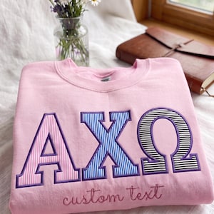May include: A light pink crewneck sweatshirt featuring large embroidered Greek letters Alpha, Chi, and Omega. Each letter has a purple border and a distinct striped pattern in pink, blue, or black and white. The words custom text are embroidered below.