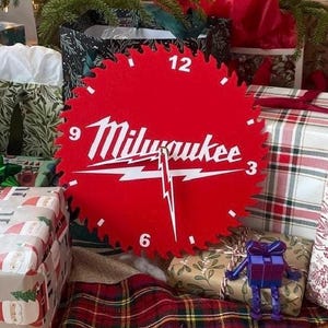 May include: A red, circular clock with a saw blade edge, featuring the "Milwaukee" logo in white. The clock has white numbers at the 3, 6, 9, and 12 positions. The clock is surrounded by wrapped gifts and Christmas decorations.