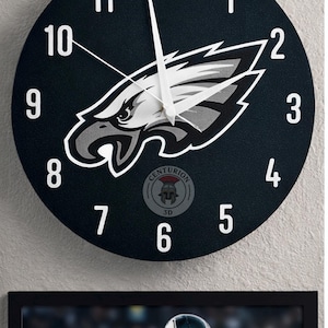 May include: A round black wall clock featuring the Philadelphia Eagles logo in white and gray. White numerals mark the hours. The clock has white hands and the text "CENTURION 3D" at the bottom. Below the clock is a framed image of a football player.