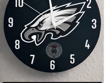 May include: A round black wall clock featuring the Philadelphia Eagles logo in white and gray. White numerals mark the hours. The clock has white hands and the text "CENTURION 3D" at the bottom. Below the clock is a framed image of a football player.