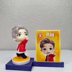 May include: A figurine of a person in a red jacket and black trousers, standing on a yellow and white base, with a matching card that says "RM". The figurine is on a blue base.