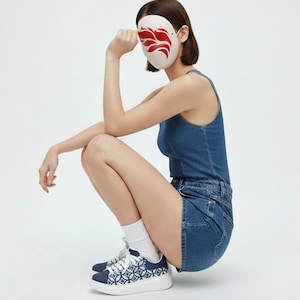 May include: A white mask with a red flame design is held up. The person is wearing a denim tank top and denim shorts. The person is also wearing white socks and blue and white patterned sneakers.