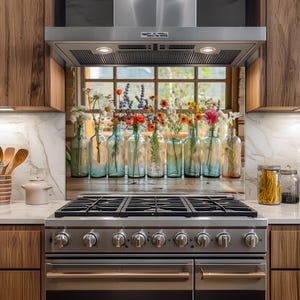 Op de afbeelding: Een keukenachterwand met een rij glazen flessen gevuld met kleurrijke wilde bloemen. De flessen zijn in verschillende tinten blauw en helder, gerangschikt voor een raam met een houten frame. De bloemen omvatten madeliefjes, rozen en andere bloemen.