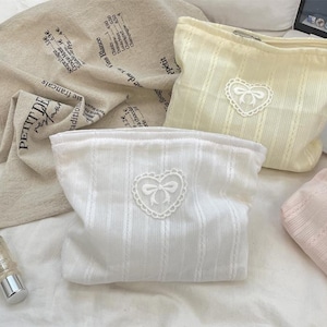 May include: Several cosmetic bags in soft pastel colors are displayed. The bags have a textured, vertical striped pattern and a heart-shaped appliqué with a bow. One bag is white, another is pale yellow, and a third is light pink.
