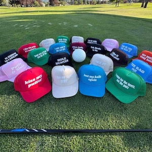 Könnte beinhalten: Eine Sammlung von Golfkappen in verschiedenen Farben, darunter Rot, Blau, Grün und Rosa, mit humorvollen Sprüchen wie "Kiss my putter" und "Lost my balls again". Ein Golfball ist ebenfalls vorhanden.