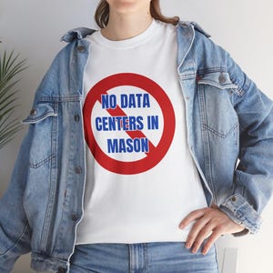 May include: White t-shirt with a red circle and a diagonal red line through it. The text inside the circle reads "NO DATA CENTERS IN MASON" in blue. The person is wearing a light blue denim jacket.