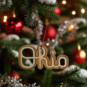 May include: Wooden Christmas ornament spelling out "Ohio" in a layered design. The ornament is light brown with a lighter wood inlay, hanging from a red string. The background features a decorated Christmas tree with red ornaments and lights.