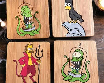 May include: Four square bamboo coasters featuring cartoon illustrations. Two coasters show a green alien with tentacles inside a glass dome. One coaster depicts a black bird with a yellow beak on a gray stone. One coaster shows a yellow devil character.