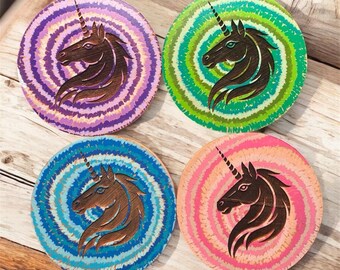 May include: Four round wooden coasters with a spiral design and a carved unicorn head. The coasters feature a variety of colors, including purple, green, blue, and pink. The unicorn is brown.