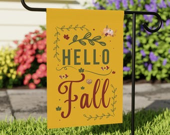 May include: A rectangular yellow garden flag hanging on a black metal stand. The flag features the text "HELLO Fall" in dark green and maroon script. Decorative elements include small illustrations of autumn leaves, a pumpkin, and a pie.