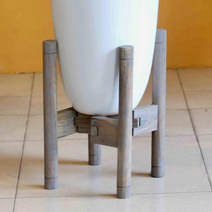 May include: A tall, white ceramic planter with a plant, set on a gray wooden stand. The planter has a smooth, glossy finish and is filled with white stones. The stand has a simple, geometric design.