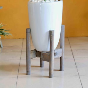 May include: A tall, white ceramic planter with a rounded shape, filled with white stones and a green plant. The planter sits on a gray wooden stand with four legs, creating a modern, minimalist aesthetic. The stand is approximately 24 inches tall.