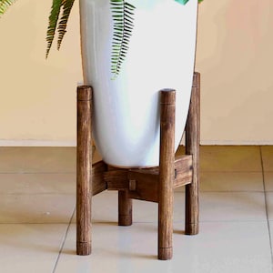 May include: A tall, white ceramic planter filled with green plants, set on a dark brown wooden plant stand. The stand has a geometric design with four vertical legs and a cross-braced base, creating a modern, minimalist aesthetic.
