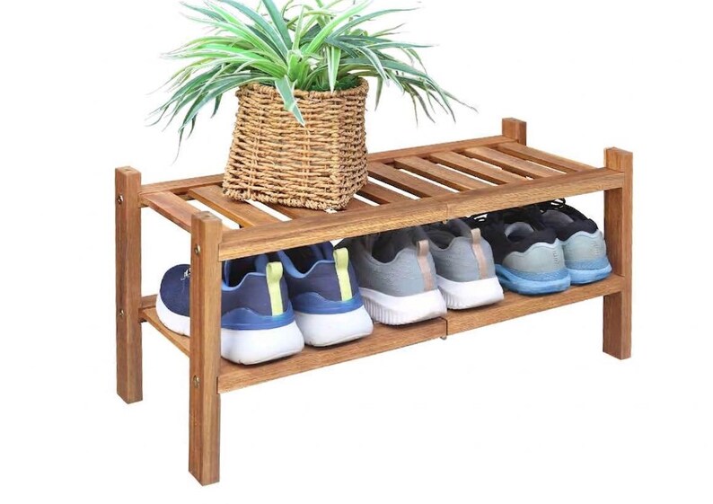 Puede incluir: Un zapatero de madera de dos niveles con acabado natural. La balda superior contiene una cesta tejida con una planta verde. La balda inferior muestra varios pares de zapatillas deportivas en varios colores, incluyendo azul, gris y negro.