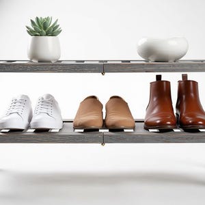 Puede incluir: Un zapatero de madera gris con dos estantes. El estante superior contiene una peque&ntilde;a suculenta en una maceta blanca y un cuenco de cer&aacute;mica blanco. El estante inferior muestra zapatillas blancas, mocasines color canela y botines de cuero marr&oacute;n.