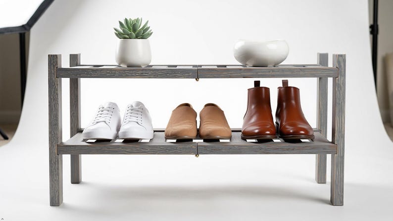 Puede incluir: Un zapatero de madera gris con dos estantes. El estante superior contiene una peque&ntilde;a suculenta en una maceta blanca y un cuenco de cer&aacute;mica blanco. El estante inferior muestra zapatillas blancas, mocasines color canela y botines de cuero marr&oacute;n.